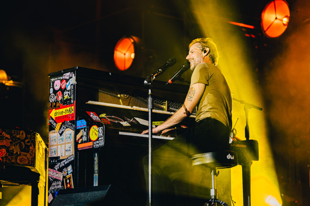 Musician playing a sticker-covered piano on a dark stage, bathed in yellow stage lights, wearing a headset microphone.