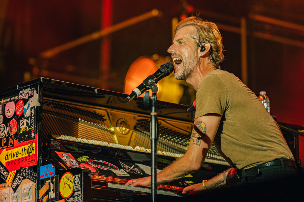 Male musician singing into a microphone while playing a sticker-covered keyboard on stage.