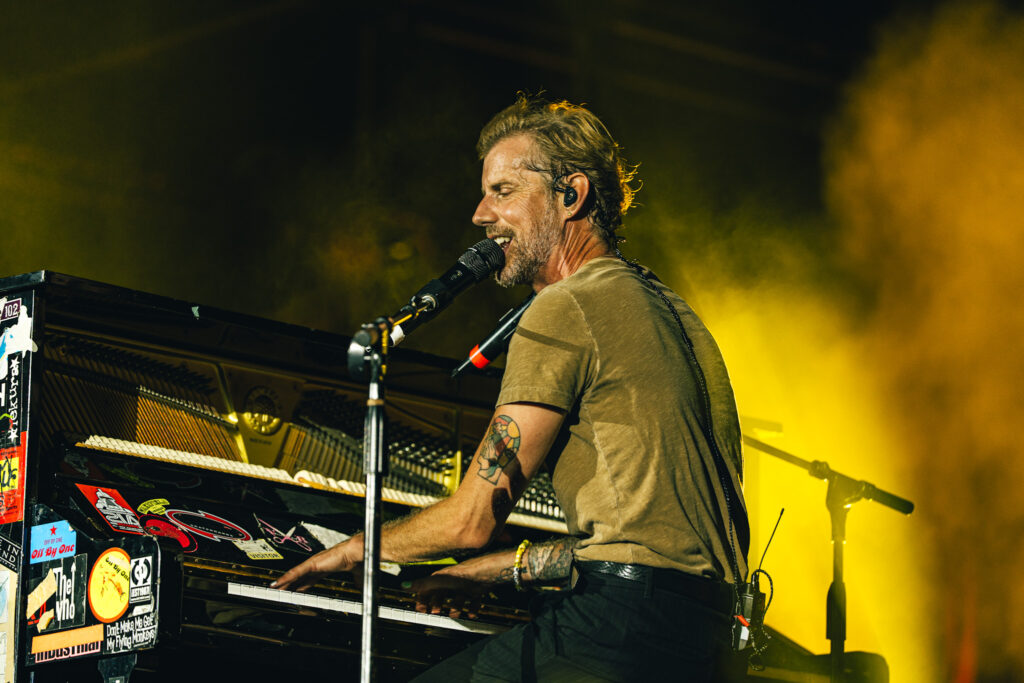 Male musician singing into a mic while playing a keyboard on stage, with yellow stage lighting and smoke in the background.