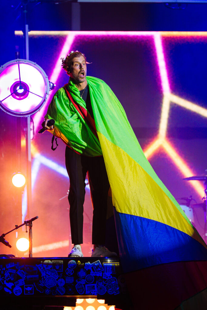 Performer on a stage wrapped in a bright rainbow flag, microphone in hand, colorful neon lights behind him.