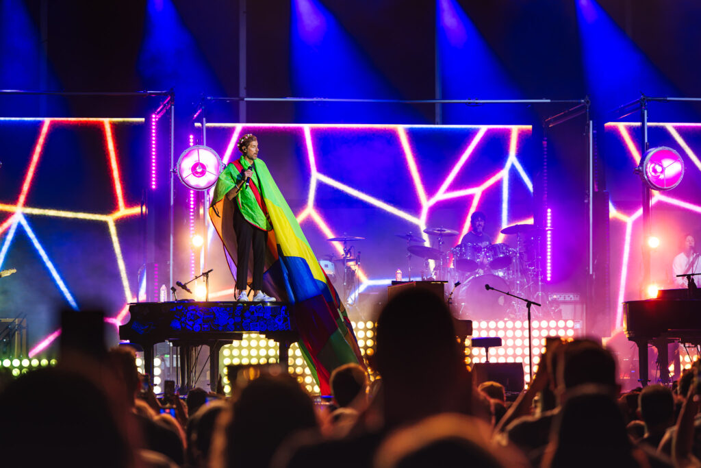 Performer on stage wearing a rainbow cape under blue and neon lights with geometric LED panels.