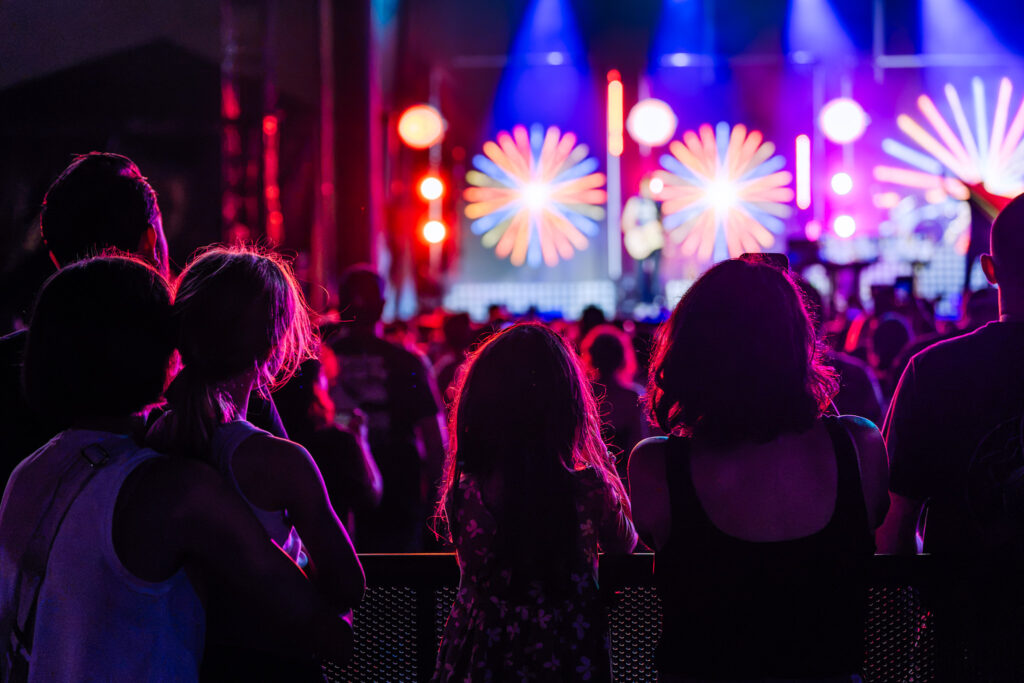 Crowd of people watching a concert with bright, colorful stage lights in red, blue, and purple hues behind them.