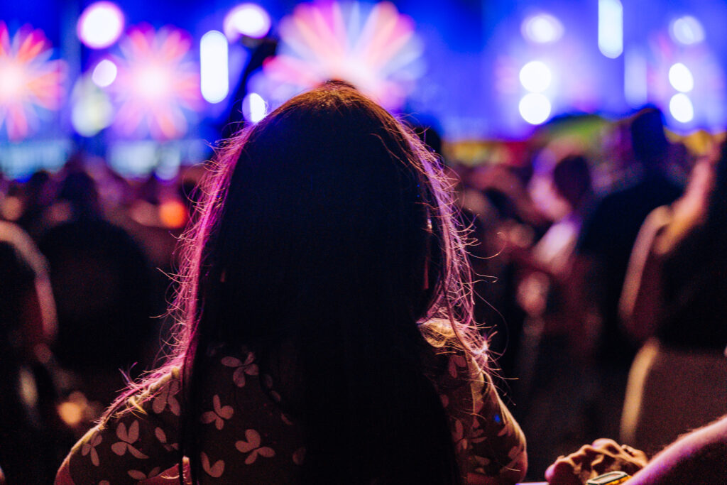 Person viewed from behind at a crowded concert, colorful stage lights glowing in the background behind their head.
