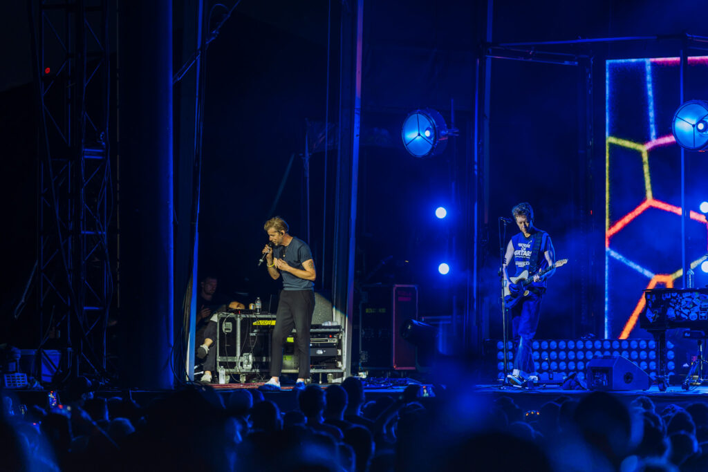 Singer with mic on a dark blue-lit stage, guitarist to the right, an energized crowd in foreground.",