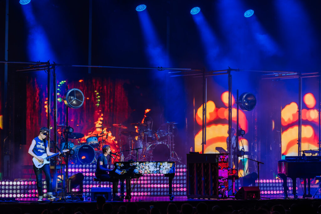 Band performs on a lit stage under blue spotlights with a colorful LED backdrop behind them, audience in shadow.