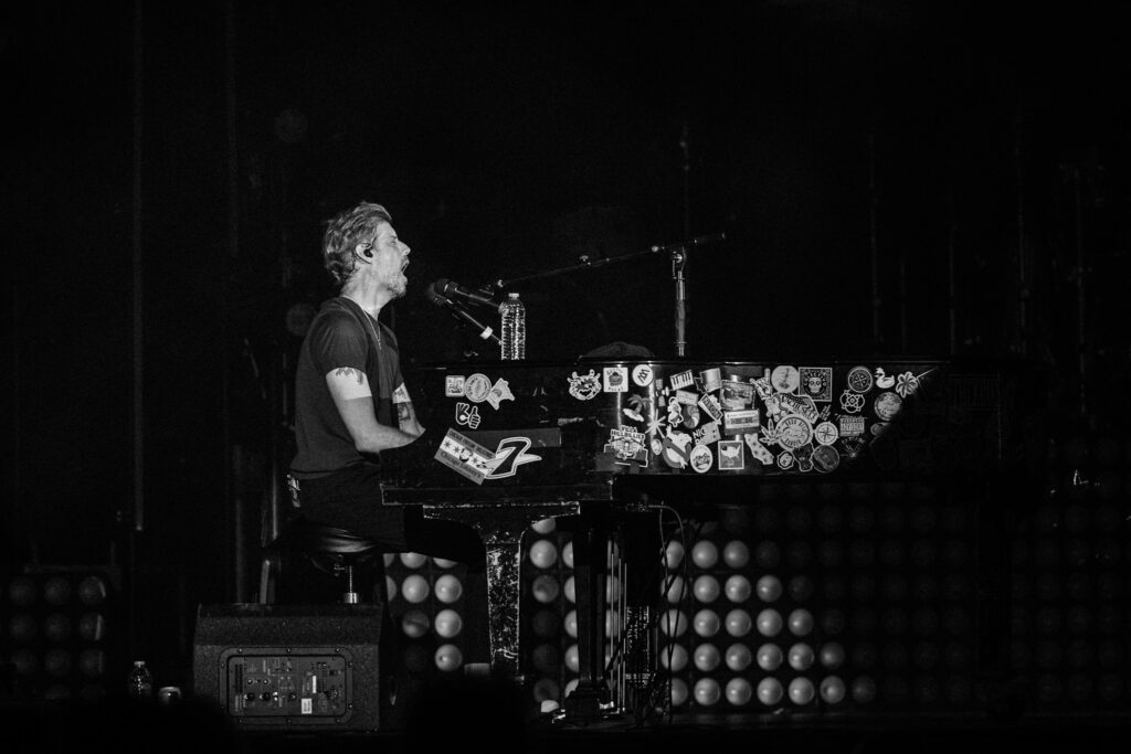 Male musician singing into a microphone while playing a sticker-covered piano on a dark stage, with a water bottle resting on the piano