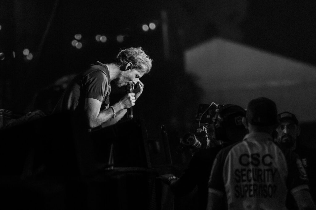 Male singer on stage, microphone in hand, leaning toward the crowd during a nighttime concert; security staff visible in the foreground.