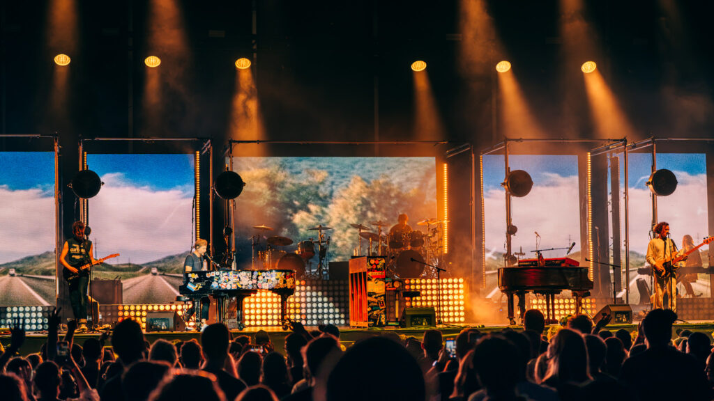 Band performs on a lit stage with multiple screens and orange spotlights, crowd waving hands in front.