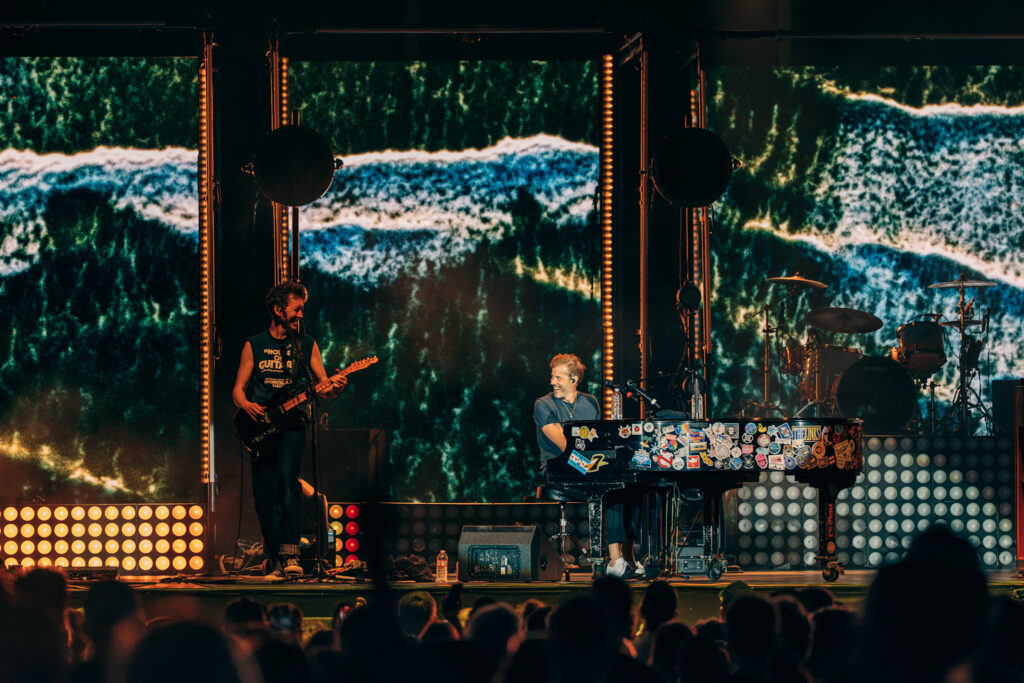 Concert stage with keyboardist at center and guitarist on the left performing, colorful LED backdrop, and drum kit on the right; audience in foreground.