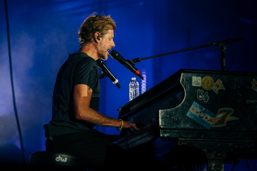 Male musician seated at a keyboard on a blue-lit stage, singing into a microphone to the left of the frame.
