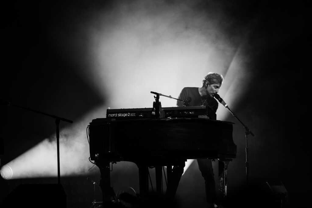 Male keyboardist performing live on a dimly lit stage with bright backlight and microphone at the keyboard spotlights behind him.
