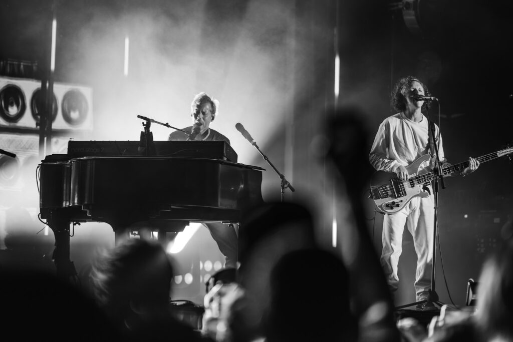 Black-and-white photo of a keyboardist at left and a bassist-singer at right performing on a smoky stage with a crowd in the foreground.
