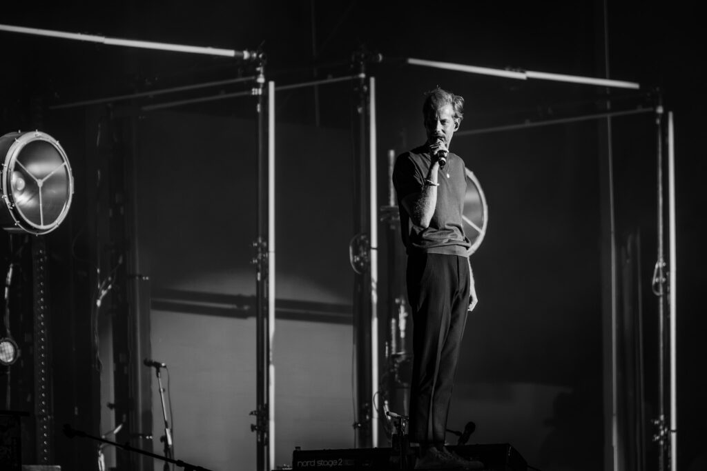 Male singer performing on a dark stage, microphone in hand, with stage equipment in the background (black & white).