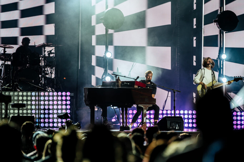 Live concert scene with a drummer on the left, keyboardist center, and guitarist on the right performing on a lit stage with a bright, geometric LED backdrop and fog.
