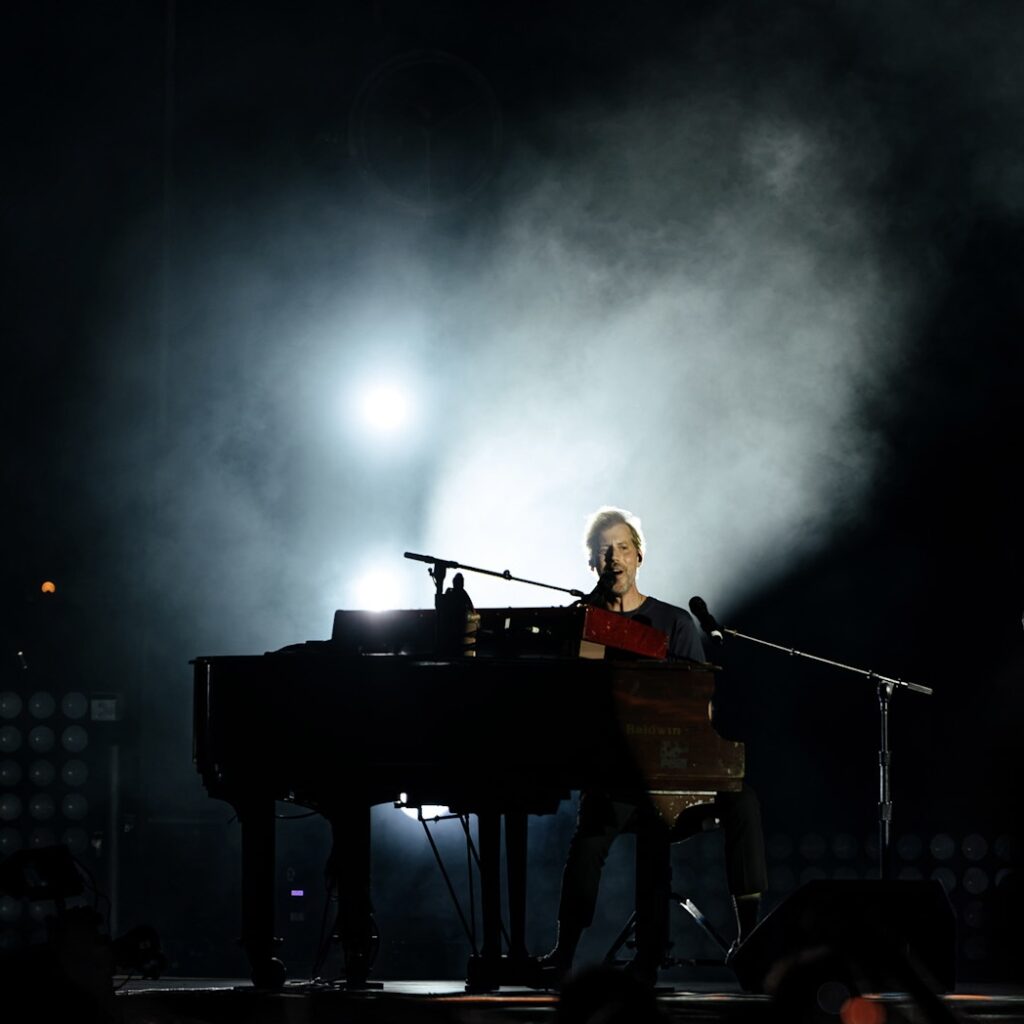 Male musician performing on a grand piano on a dark stage with smoke and backlight.