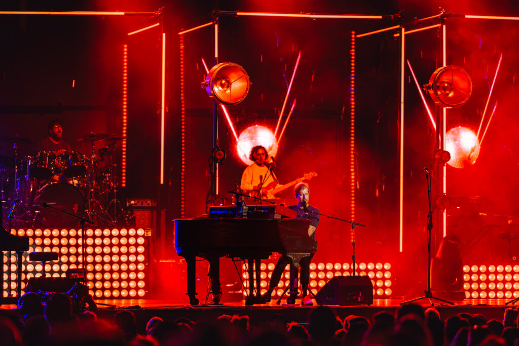 Band performs on a red-lit stage with glowing vertical LED light bars; drummer on the left, keyboardist center, audience silhouettes in front