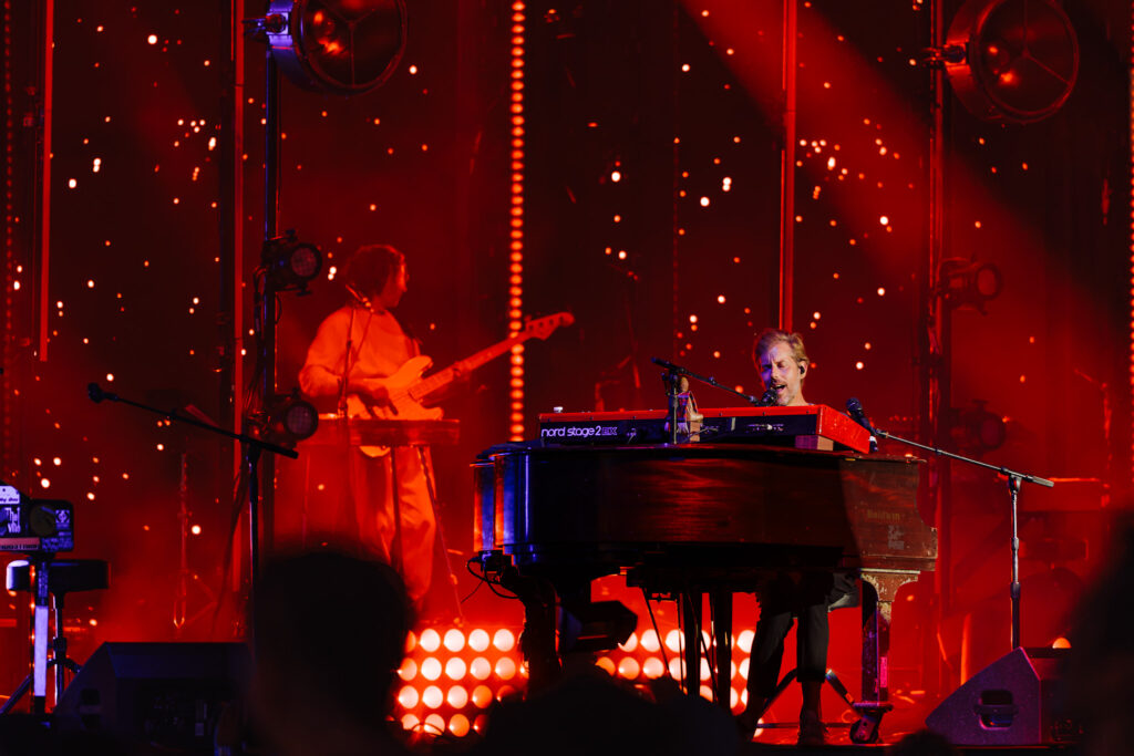 Musician playing a red Nord keyboard center stage with red lighting and a guitarist in the background
