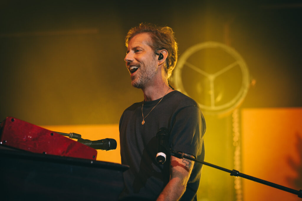 Male keyboardist mid-song on stage, singing into a microphone under warm yellow lighting.