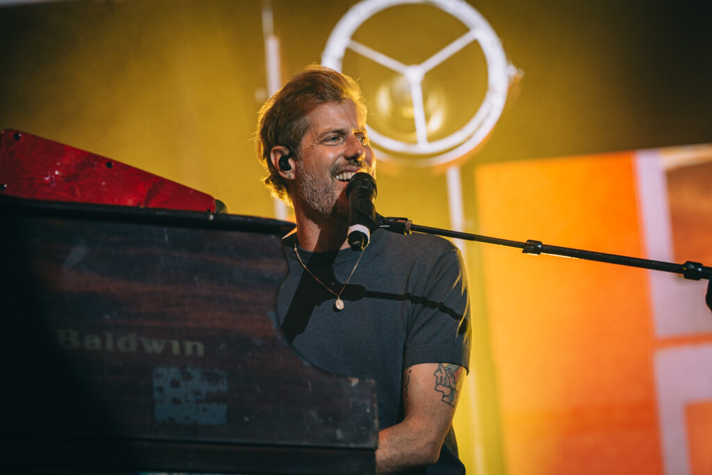 Male singer at a piano, singing into a microphone on a warmly lit stage with a circular light behind him.