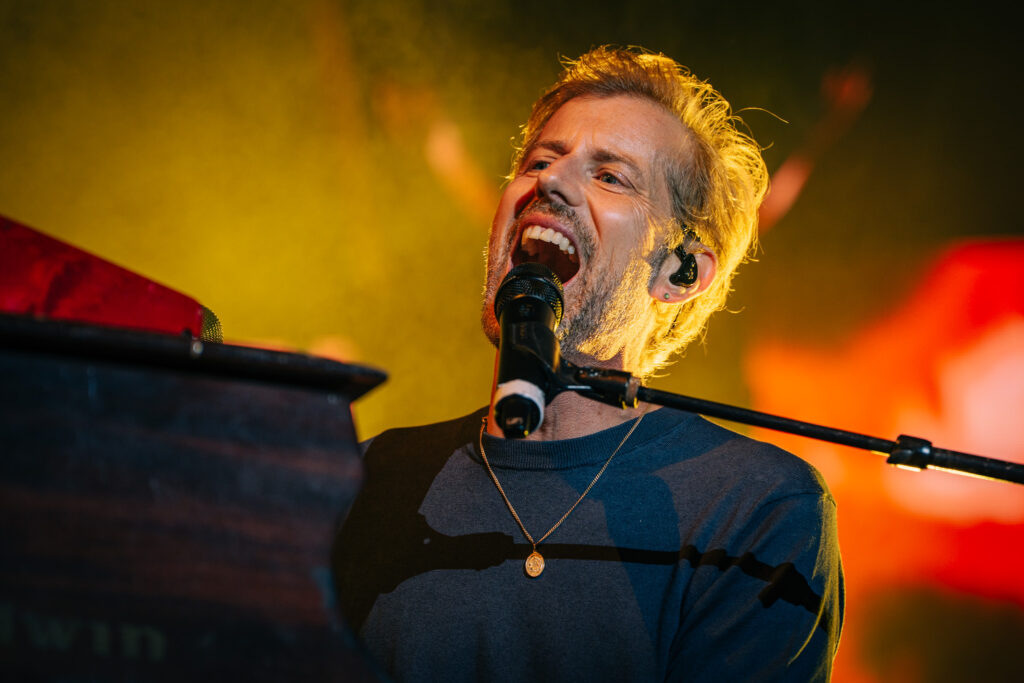 Male singer performing on stage, mouth open while singing into a microphone, wearing a necklace and blue shirt, warm orange stage lighting behind him in a live show.