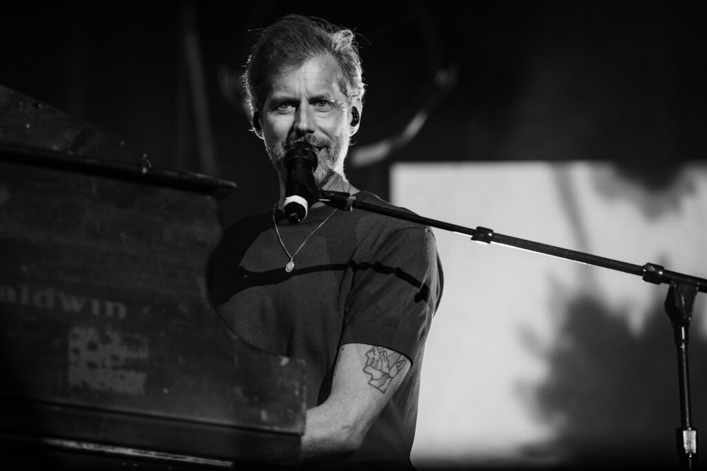 Male musician singing into a microphone at a piano during a live performance, wearing a necklace and showing a tattooed arm (black and white).