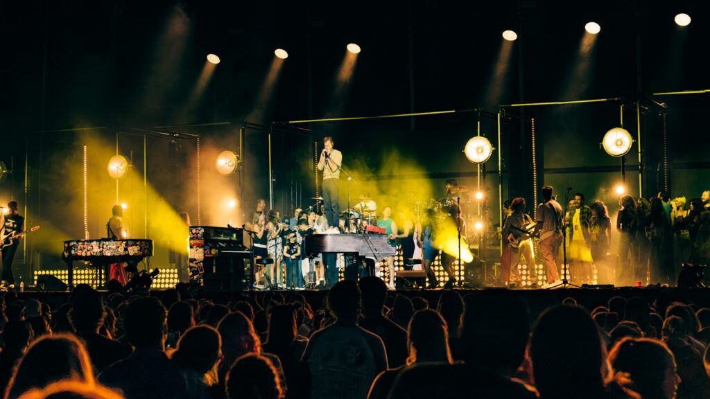 Live concert with a singer on a raised platform center stage amid yellow lighting and a cheering crowd.