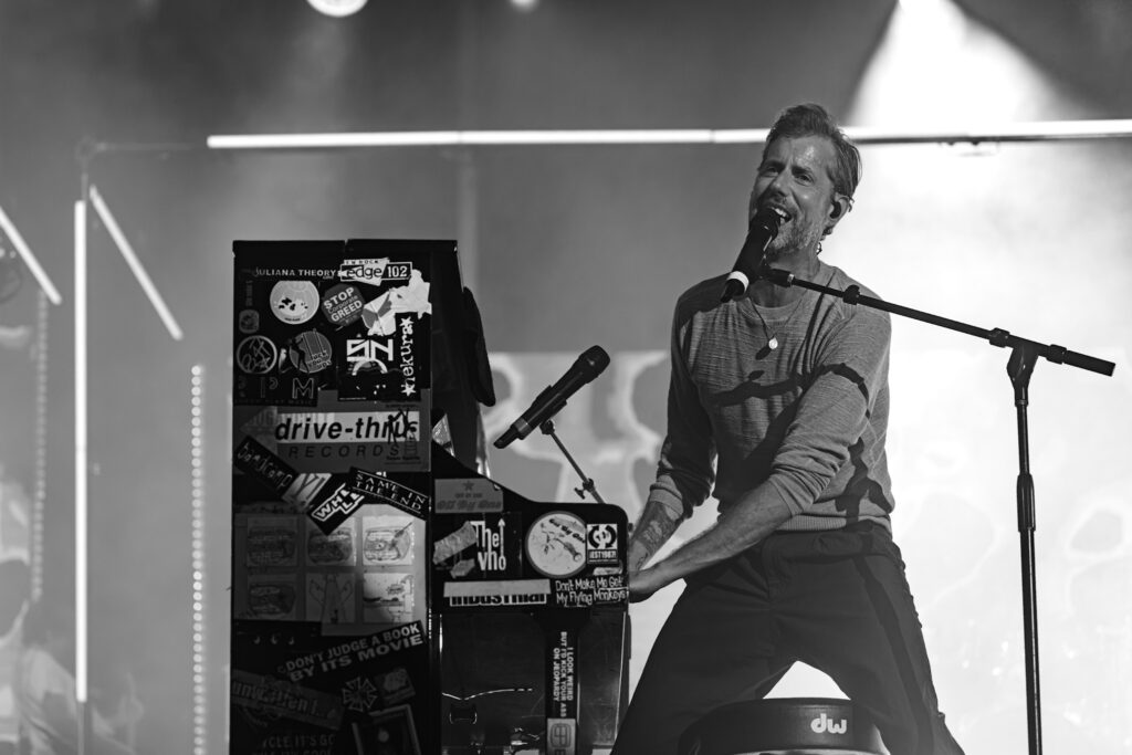 Male musician singing into a microphone at a keyboard covered in stickers on a dimly lit stage, black and white photo