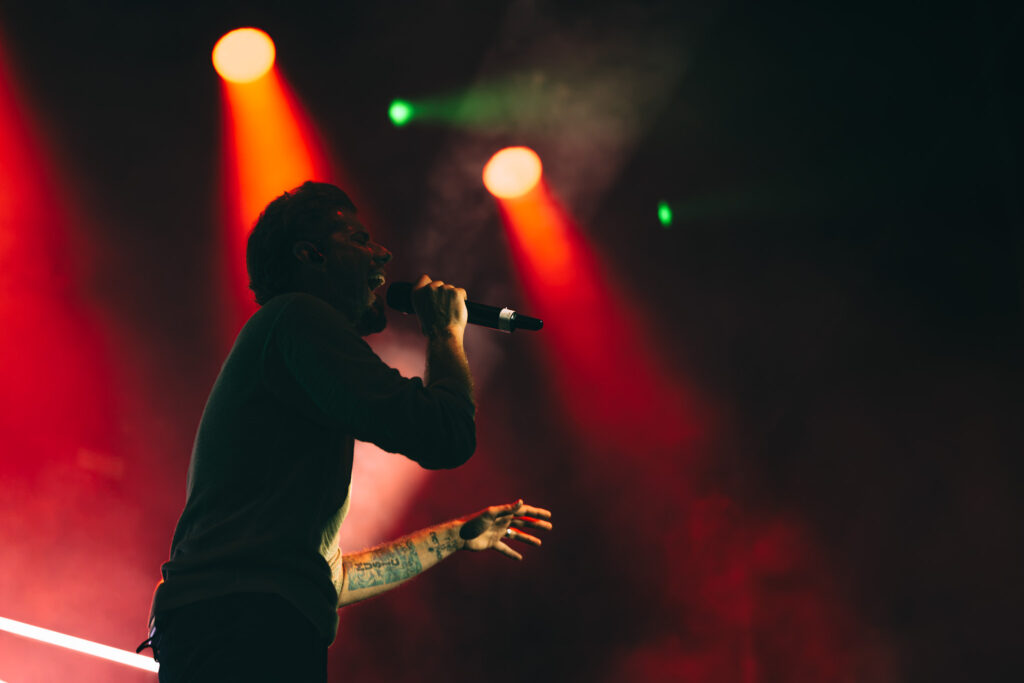 Male singer performing on stage, holding a microphone under red stage lights with a tattooed arm visible in foreground (concert scene)
