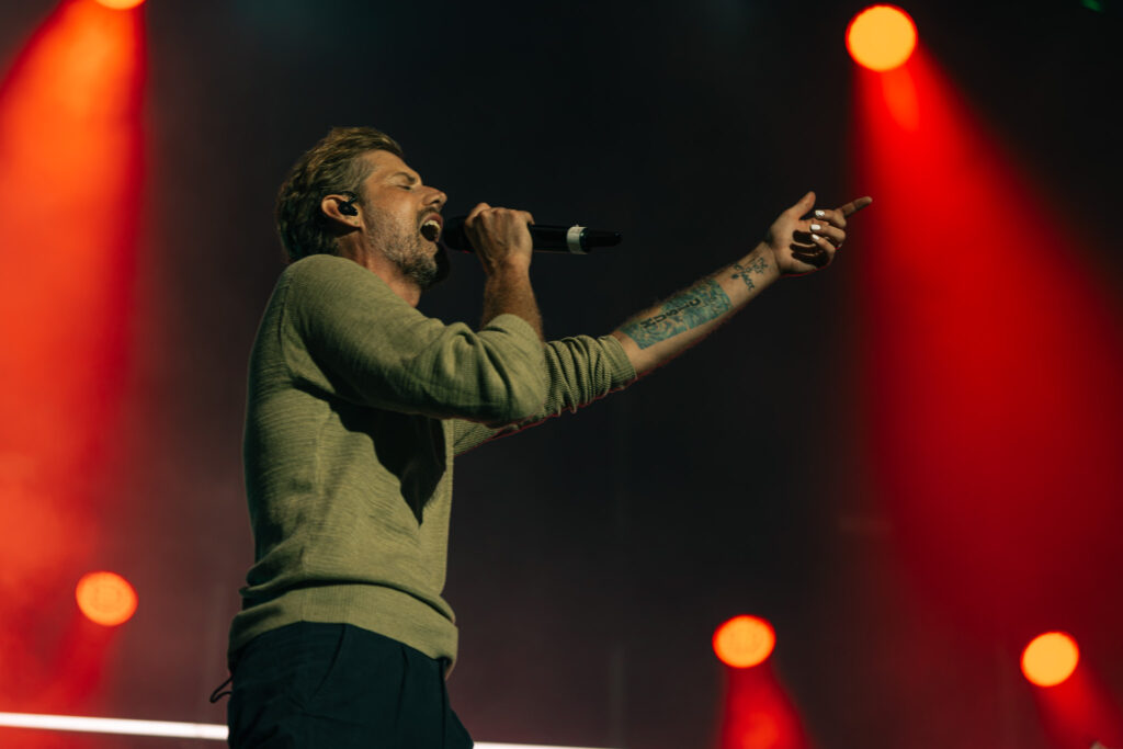 Male singer wearing a green sweater performs into a microphone on stage, red spotlights in the background.