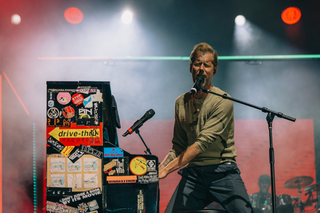 Male musician singing into a microphone while playing a sticker-covered keyboard on a smoky stage.