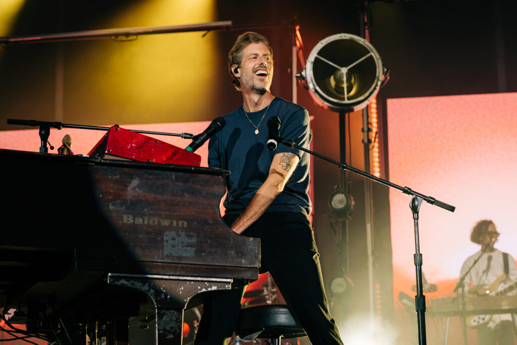 Male musician singing while playing a Baldwin keyboard on stage, wearing a dark blue shirt and a pendant necklace.