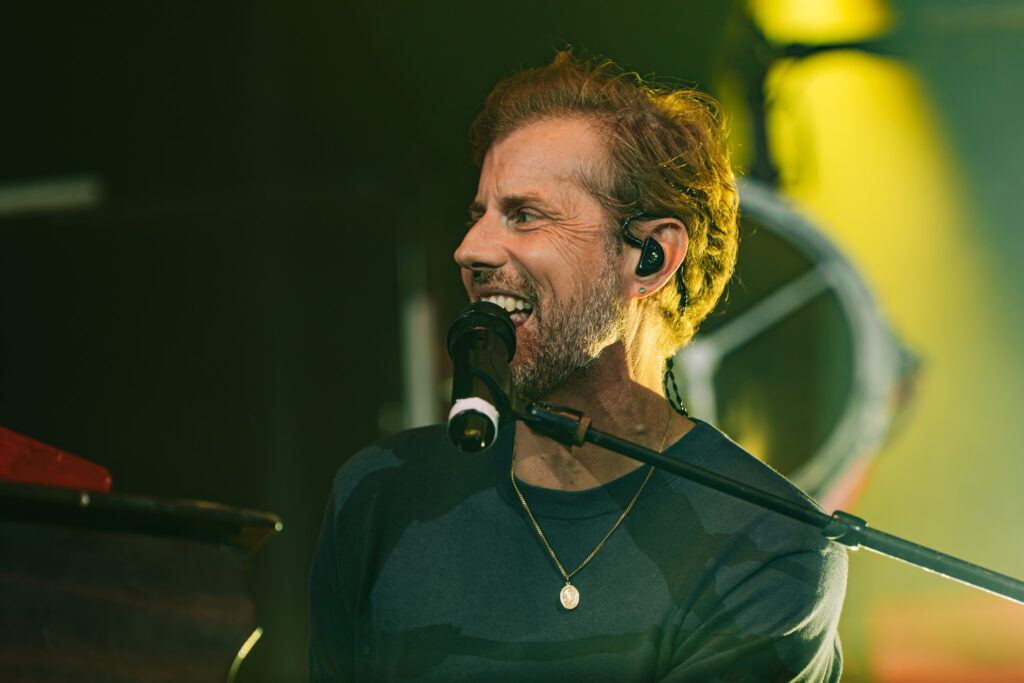 Male singer on stage, singing into a microphone with in-ear monitor, warm yellow-green stage lighting in the background.