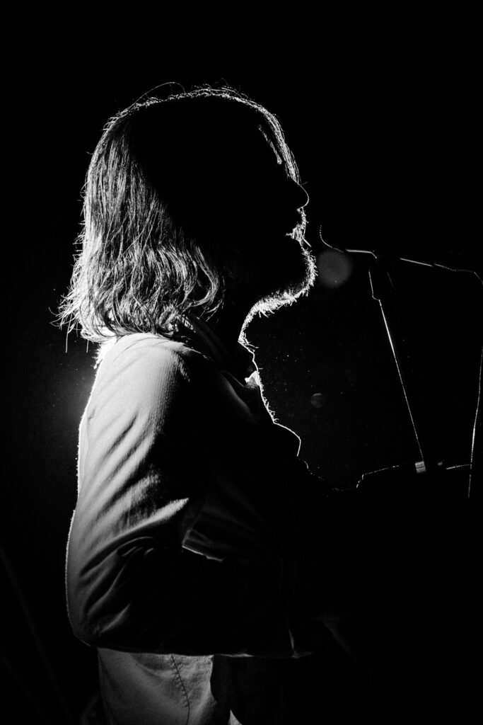 Silhouette of a bearded man singing into a microphone onstage, backlit from behind.