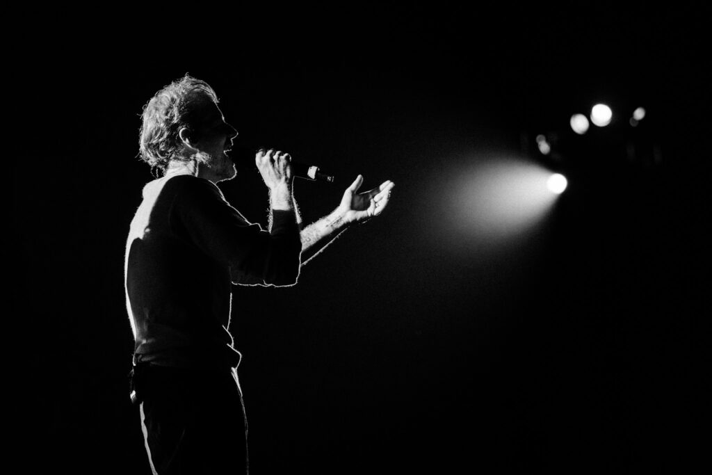 Male singer in profile singing into a handheld mic on a dark stage with a bright spotlight behind him.