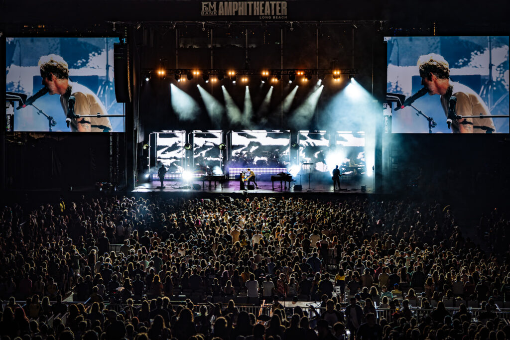 Wide view of a nighttime concert with a large stage, bright spotlights, and two giant screens showing a performer, surrounded by a packed audience.