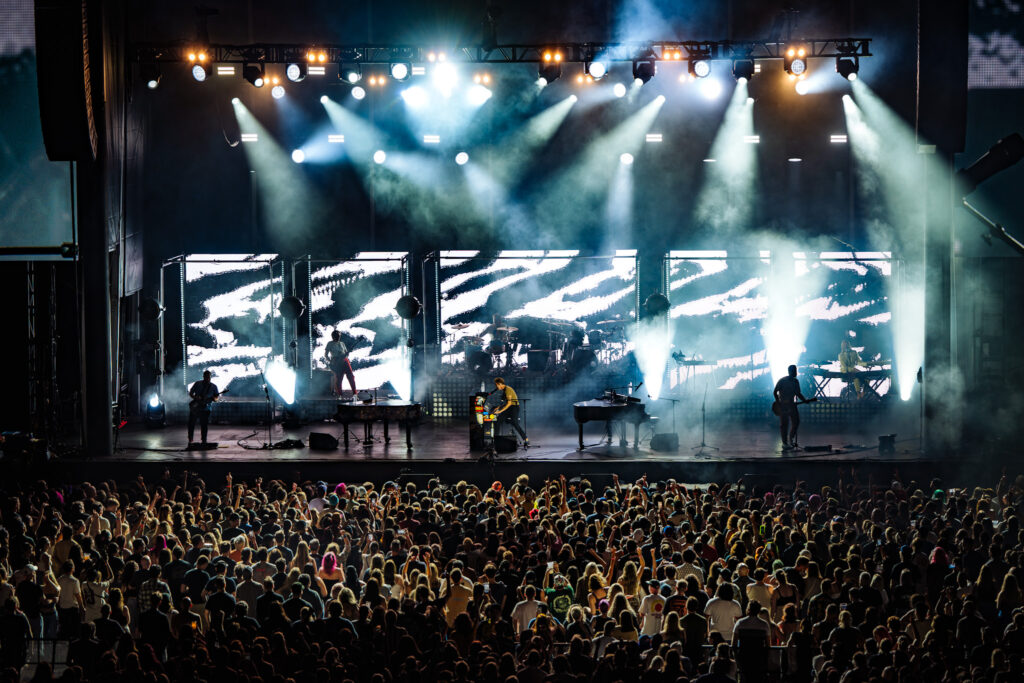 Large outdoor concert stage with bright white and colored lights, a band performing, and a cheering crowd in front.