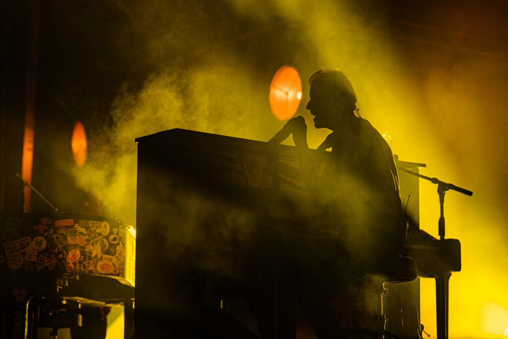 Silhouetted musician singing into a microphone at a keyboard on a smoky, yellow-lit stage.