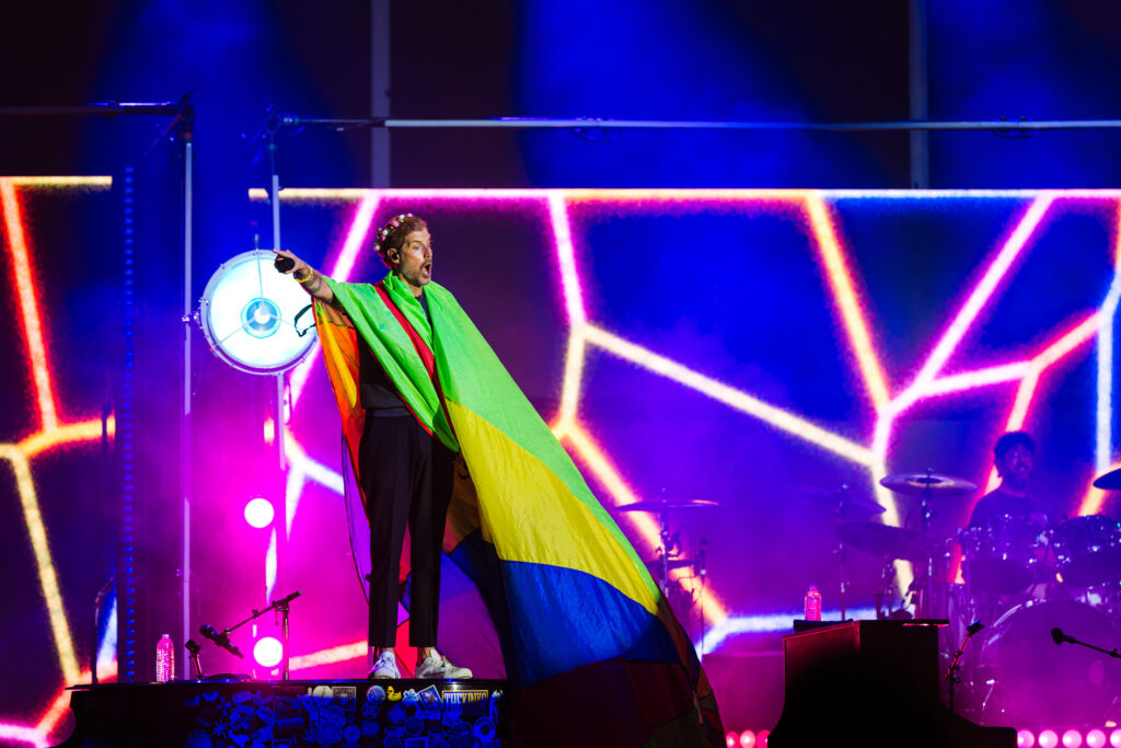 Singer on stage wrapped in a rainbow cape, holding a microphone amid bright neon lights on a colorful concert backdrop.