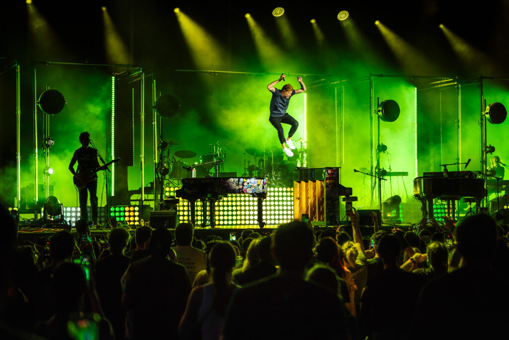 Male singer mid-air jump on a neon-lit stage with green and yellow lights, band setup, and cheering crowd.