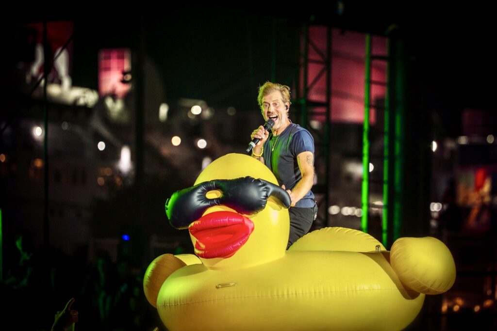 Male singer holding a microphone stands on a giant yellow inflatable duck during a nighttime concert, colorful stage lights behind.