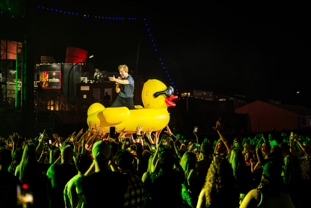 Performer singing on a large yellow inflatable duck amid a crowded night-time concert scene.