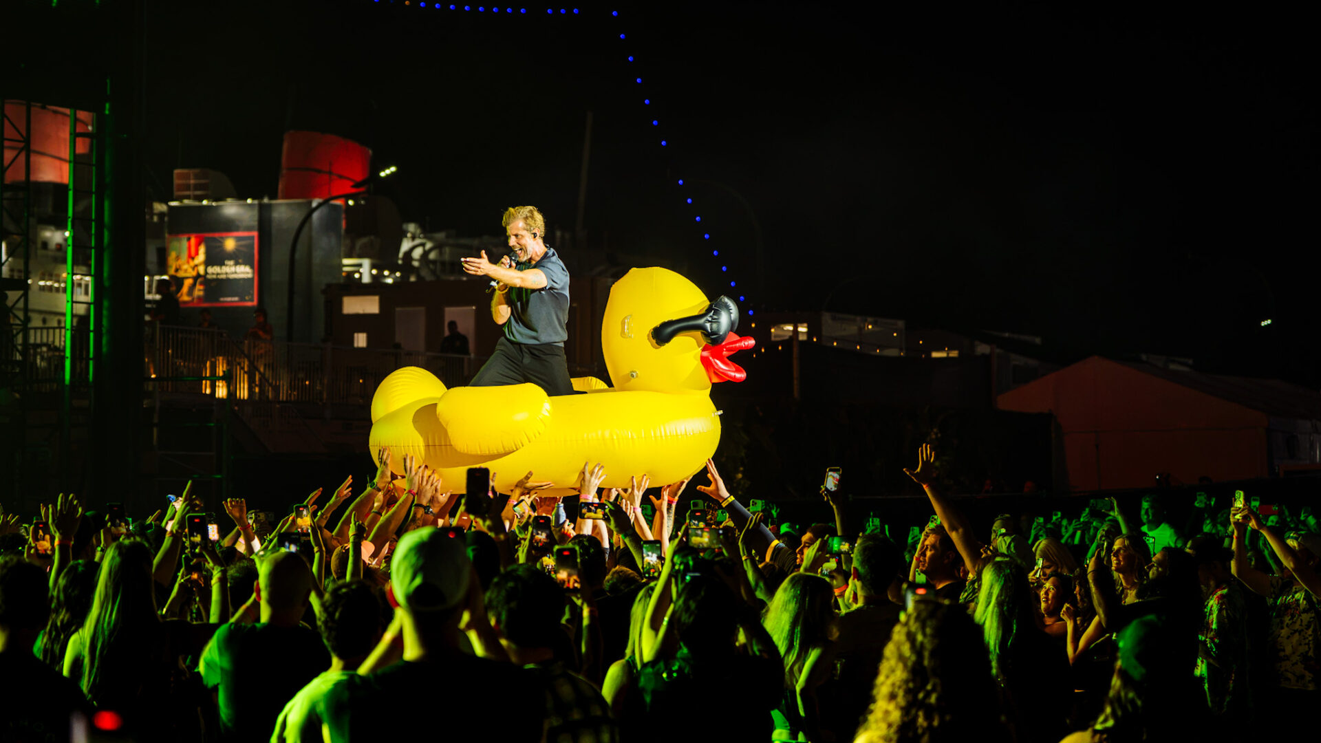 Performer singing on a large yellow inflatable duck amid a crowded night-time concert scene.