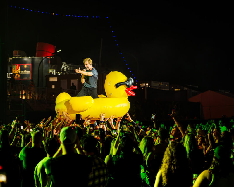 Performer singing on a large yellow inflatable duck amid a crowded night-time concert scene.