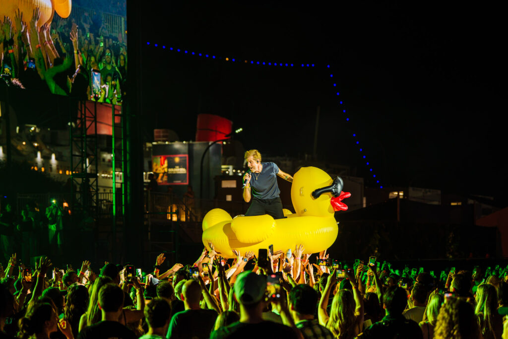 Performer with microphone atop a giant yellow inflatable duck at night, crowd raising hands around the stage.