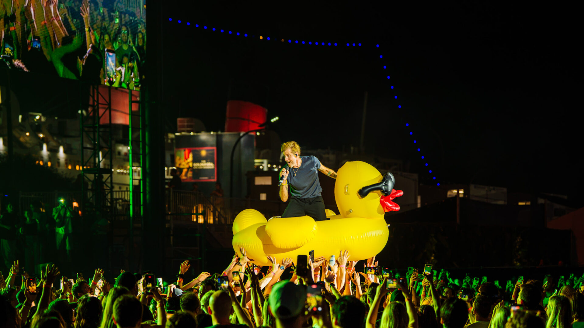 Performer with microphone atop a giant yellow inflatable duck at night, crowd raising hands around the stage.