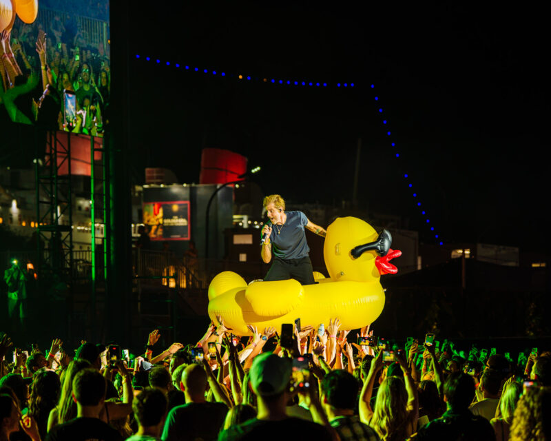 Performer with microphone atop a giant yellow inflatable duck at night, crowd raising hands around the stage.