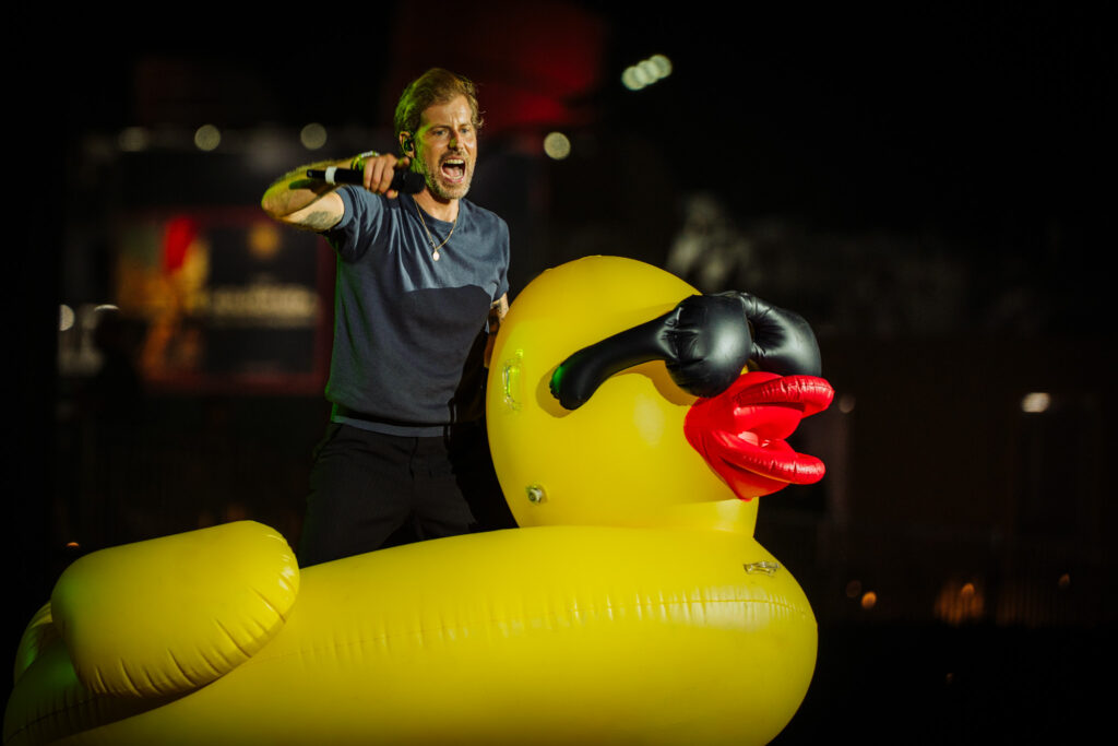 Male singer holding a microphone on stage at night, standing inside a large yellow inflatable duck structure with a red beak.