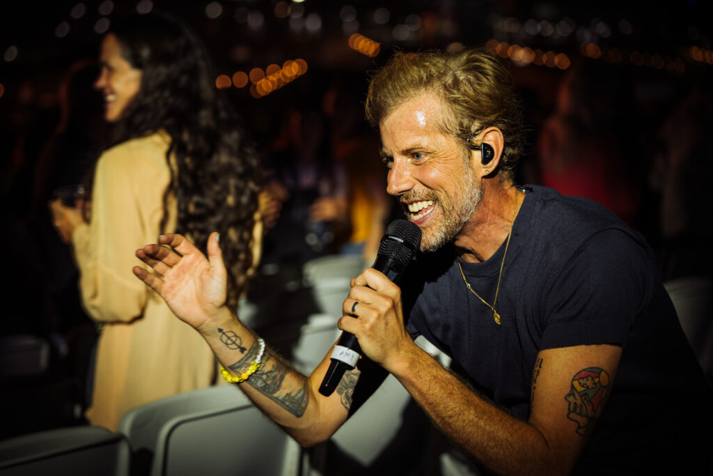 Man singing into a handheld microphone at a nighttime party, wearing an in-ear monitor and gold necklace, tattoos on his arm visible.