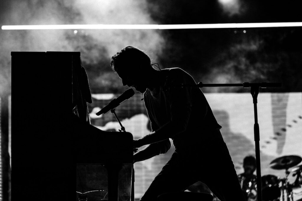 Silhouetted musician at a keyboard singing into a microphone on stage, with smoky lighting in the background.