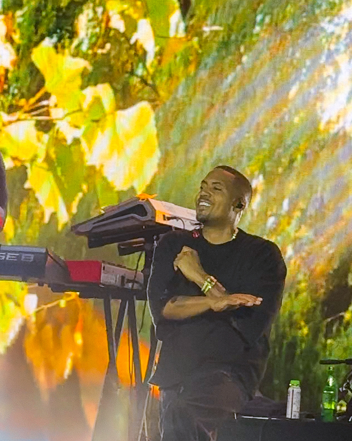 Screenshot – The 824 Journal Smiling Black male musician on stage with a keyboard behind him outdoors, arms crossed, performing joyfully.